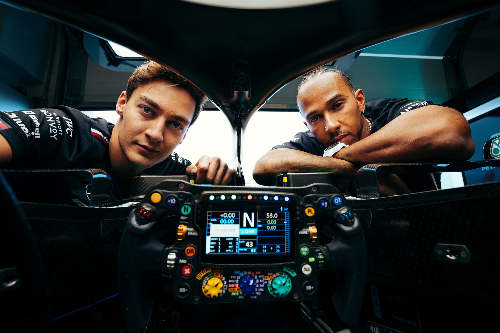 Two Formula racing drivers lean over an open cockpit with a steering wheel and digital dashboard visible inside a high-performance race car garage.