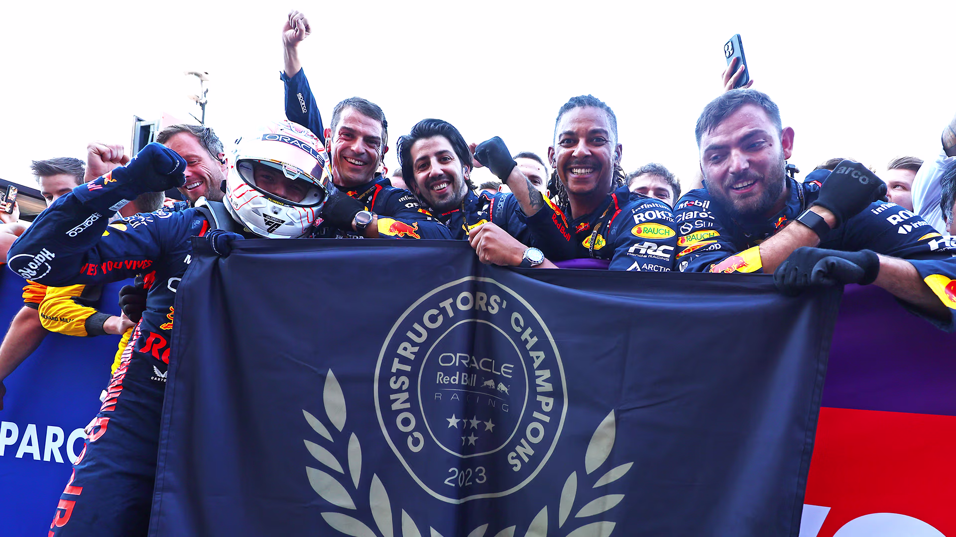 Max Verstappen and mechanics of the 2023 Red Bull F1 Team celebrating behind a Constructors' Champions 2023 flag.