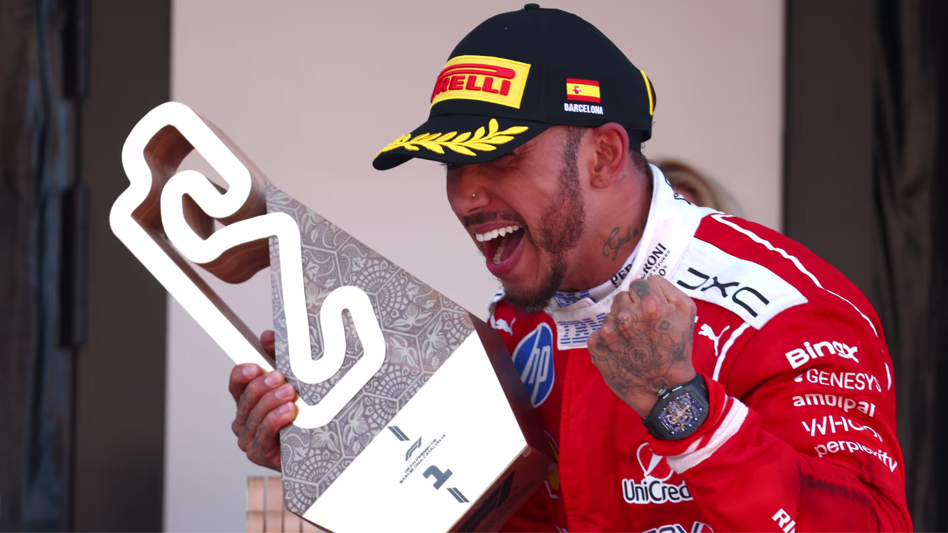 Lewis Hamilton in a red Ferrari race suit cheering and holding his first-place trophy at the 2026 Barcelona-Catalunya Grand Prix.